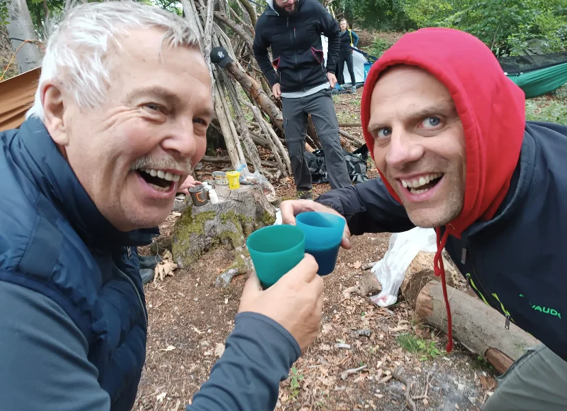 Zwei Männer stoßen mit bunten Campingbechern an und lachen fröhlich. Im Hintergrund sind Zelte, Rucksäcke und eine Gruppe von Menschen im Wald zu sehen. | © DAV Dortmund