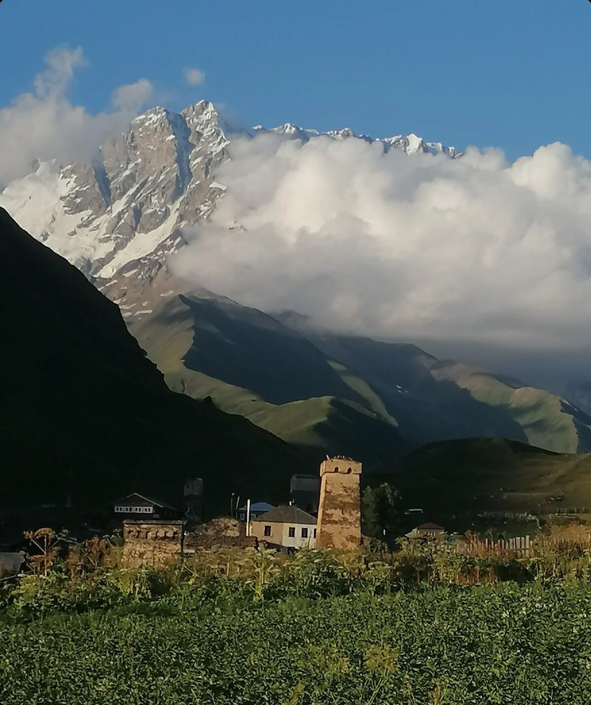 Ein kleines Dorf mit alten Steintürmen vor imposanten Bergen, teils von Wolken verdeckt, in grüner Landschaft | © DAV Dortmund