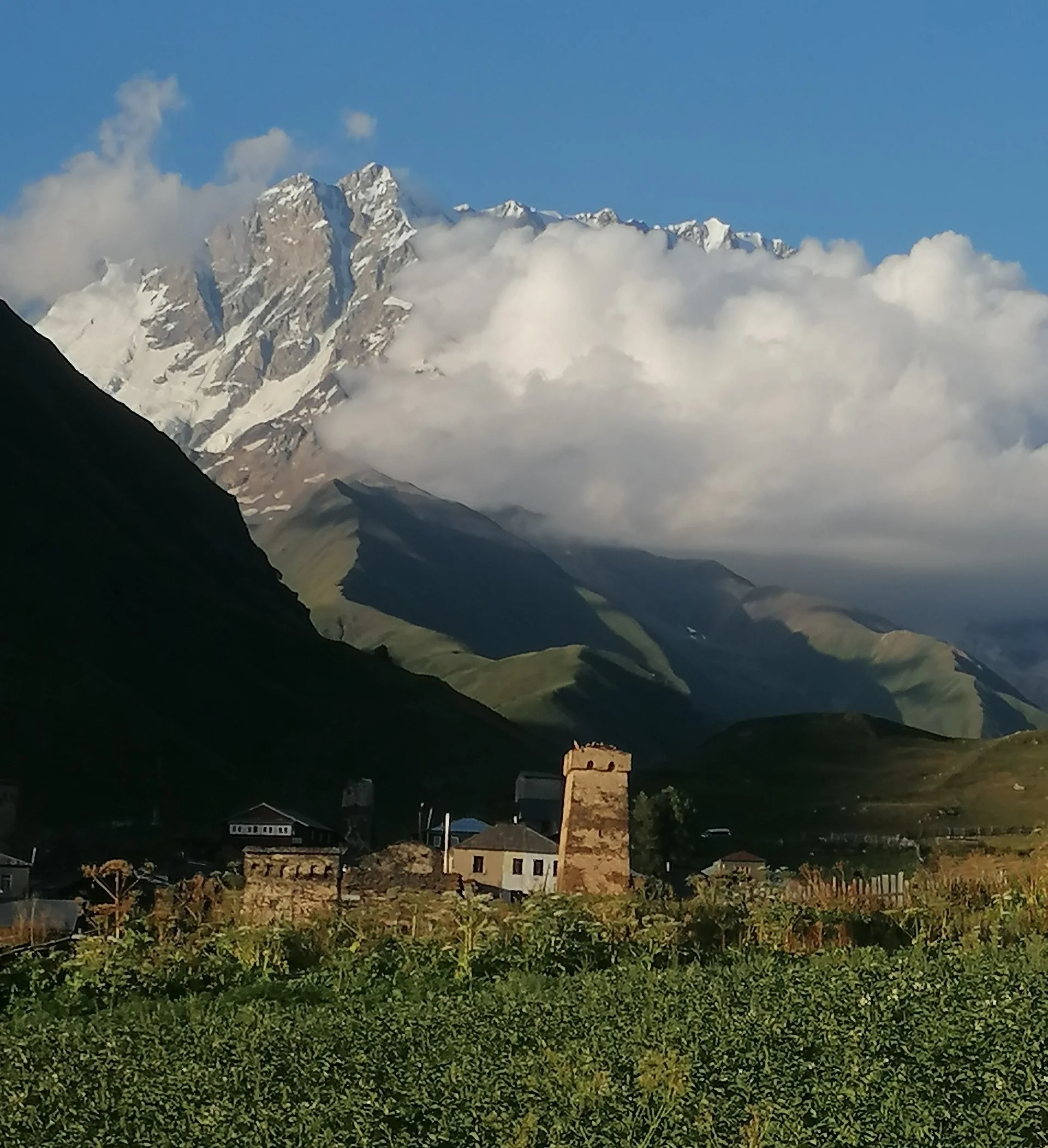 Ein kleines Dorf mit alten Steintürmen vor imposanten Bergen, teils von Wolken verdeckt, in grüner Landschaft | © DAV Dortmund