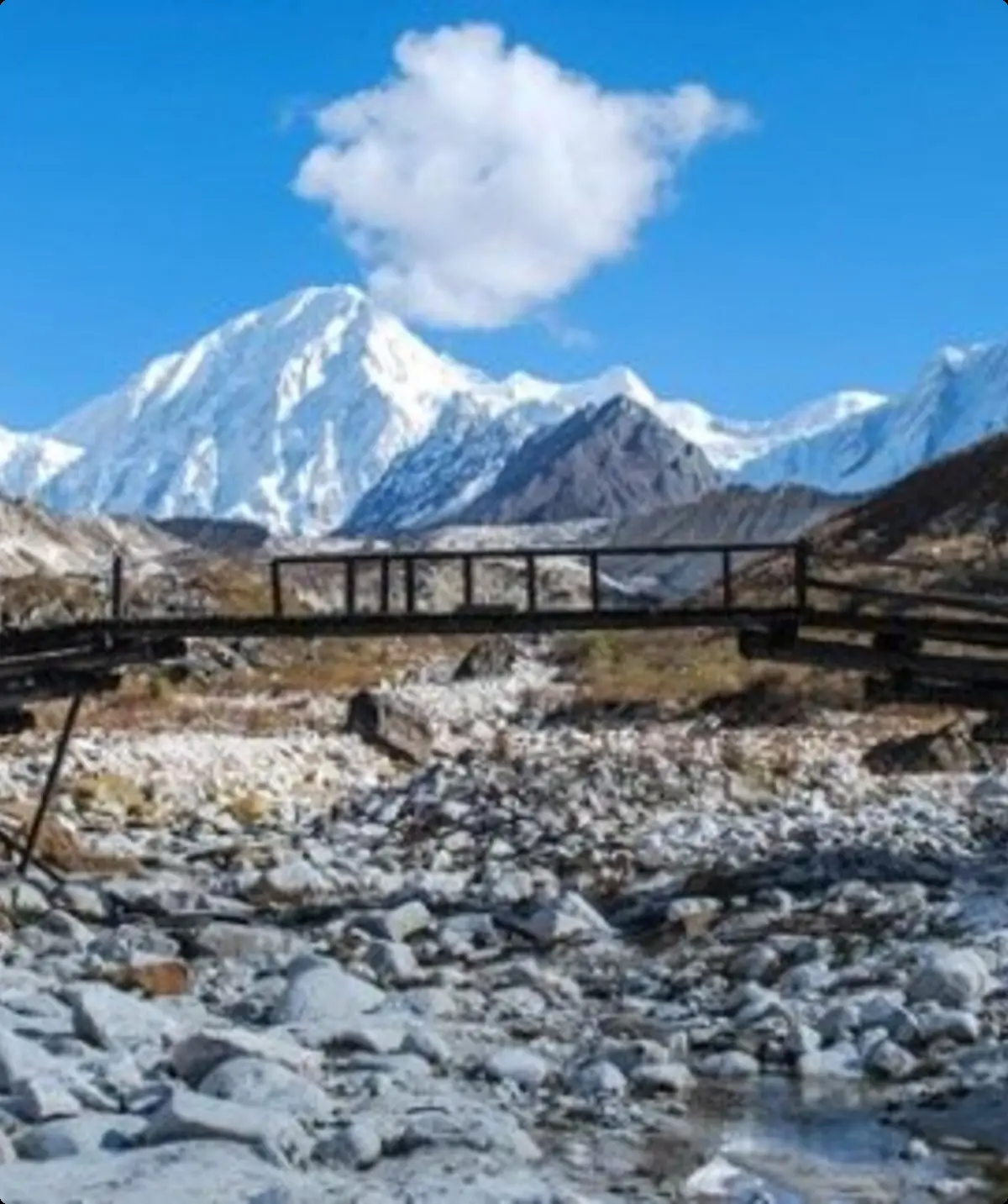 Eine beschädigte Holzbrücke überspannt ein steiniges Flussbett, im Hintergrund schneebedeckte Berge unter blauem Himmel | © DAV Dortmund