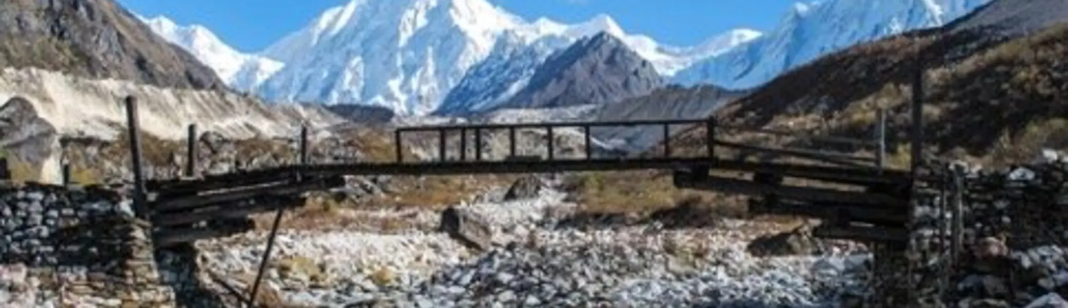 Eine beschädigte Holzbrücke überspannt ein steiniges Flussbett, im Hintergrund schneebedeckte Berge unter blauem Himmel | © DAV Dortmund