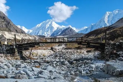 Eine beschädigte Holzbrücke überspannt ein steiniges Flussbett, im Hintergrund schneebedeckte Berge unter blauem Himmel | © DAV Dortmund