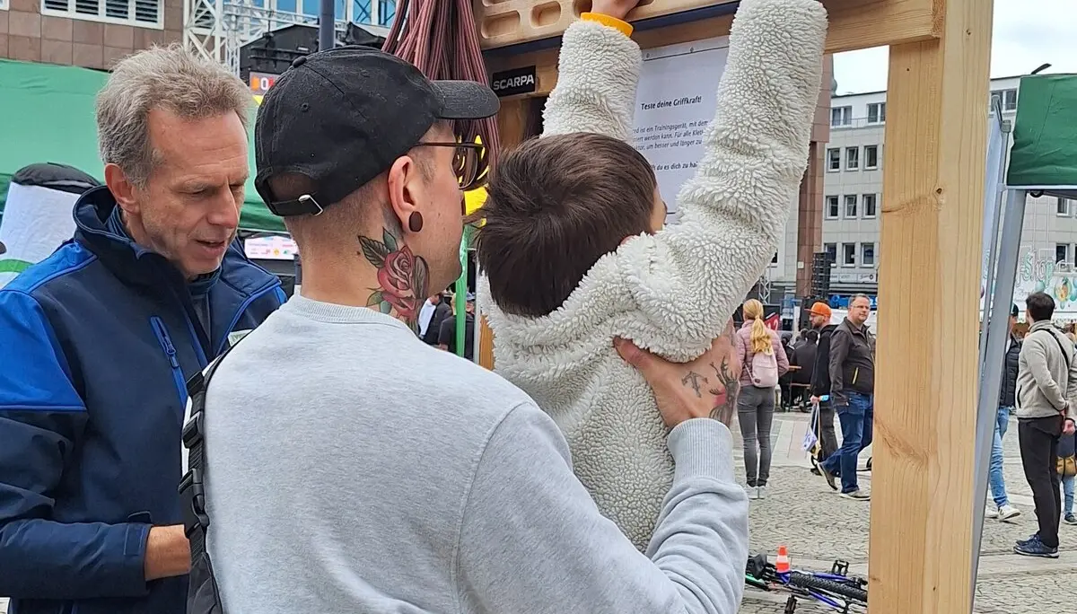 Ein Kind wird beim Hangeln an einem mobilen Trainingsboard auf einem Stadtplatz von zwei Erwachsenen unterstützt. | © DAV Dortmund