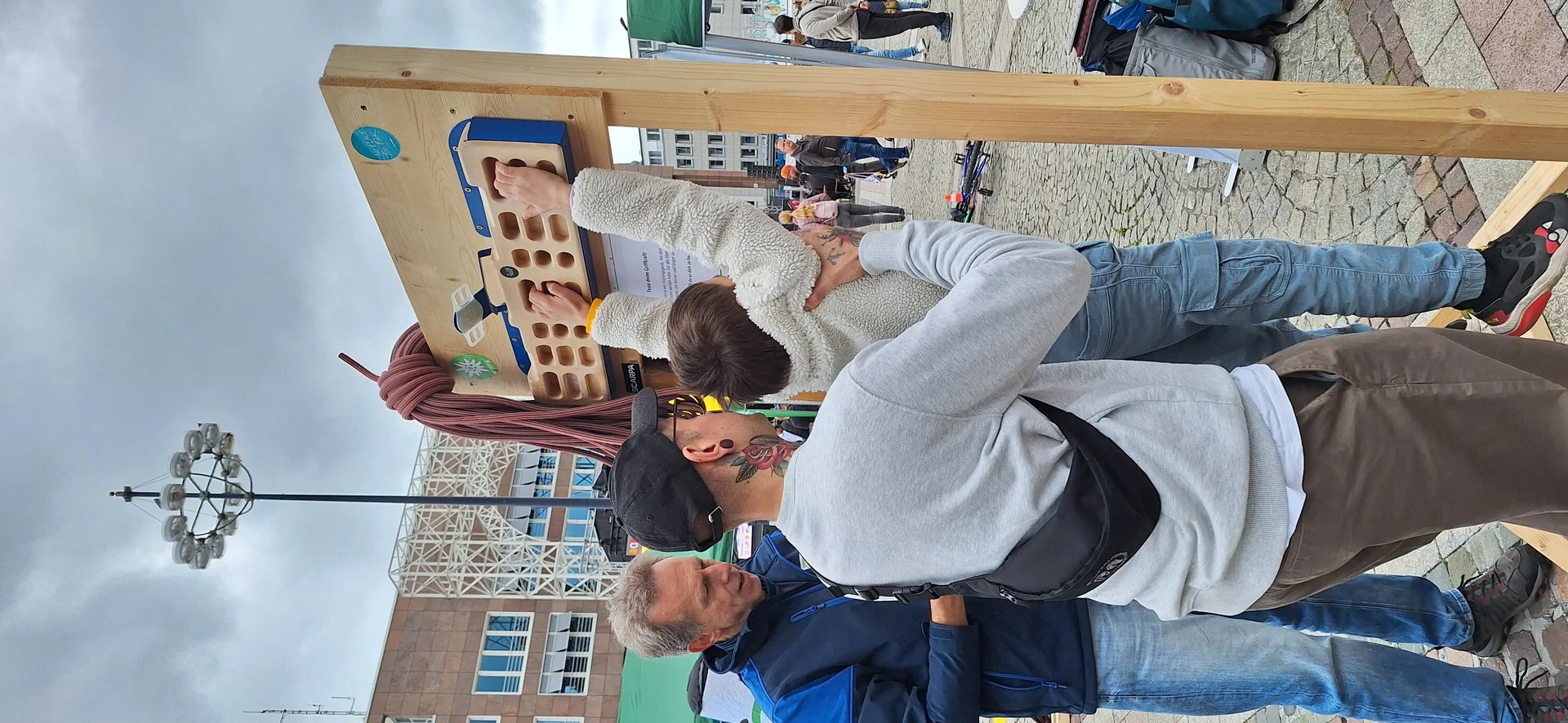 Ein Kind wird beim Hangeln an einem mobilen Trainingsboard auf einem Stadtplatz von zwei Erwachsenen unterstützt. | © DAV Dortmund