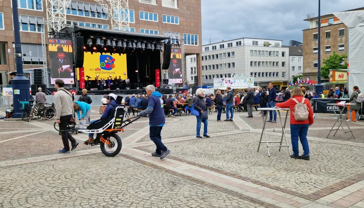 Person wird mit einer Joëlette über den Platz gefahren, im Hintergrund Bühne und Zuschauer*innen. | © DAV Dortmund