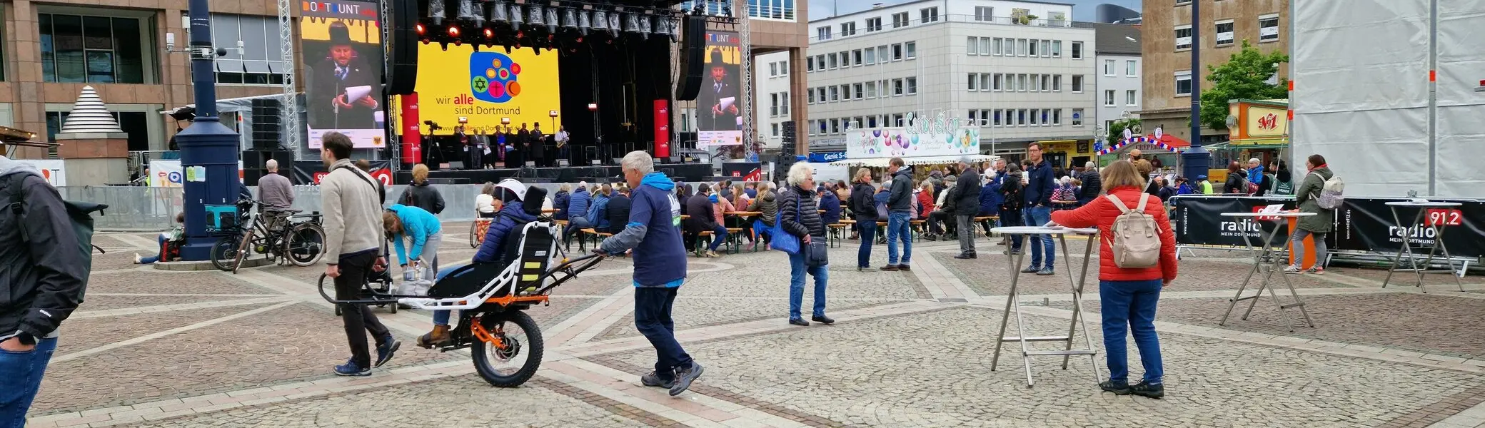 Person wird mit einer Joëlette über den Platz gefahren, im Hintergrund Bühne und Zuschauer*innen. | © DAV Dortmund
