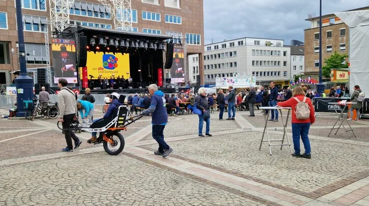 Person wird mit einer Joëlette über den Platz gefahren, im Hintergrund Bühne und Zuschauer*innen. | © DAV Dortmund