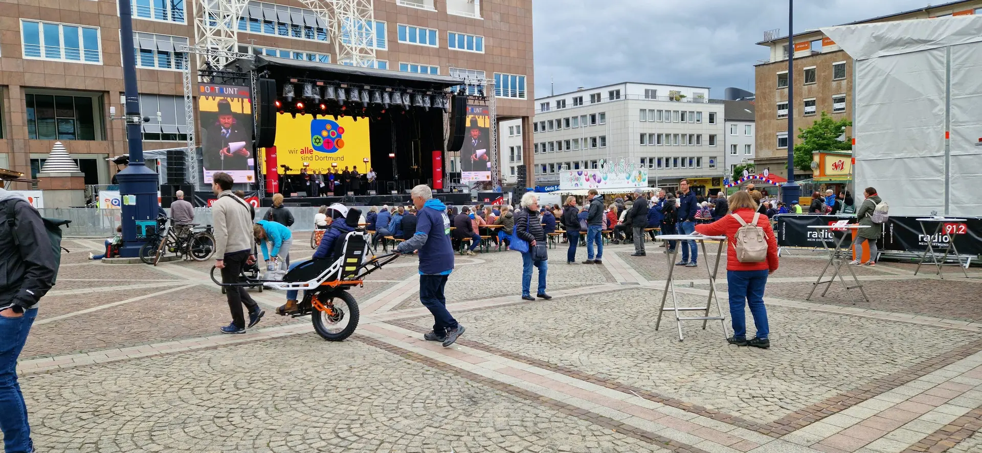 Person wird mit einer Joëlette über den Platz gefahren, im Hintergrund Bühne und Zuschauer*innen. | © DAV Dortmund