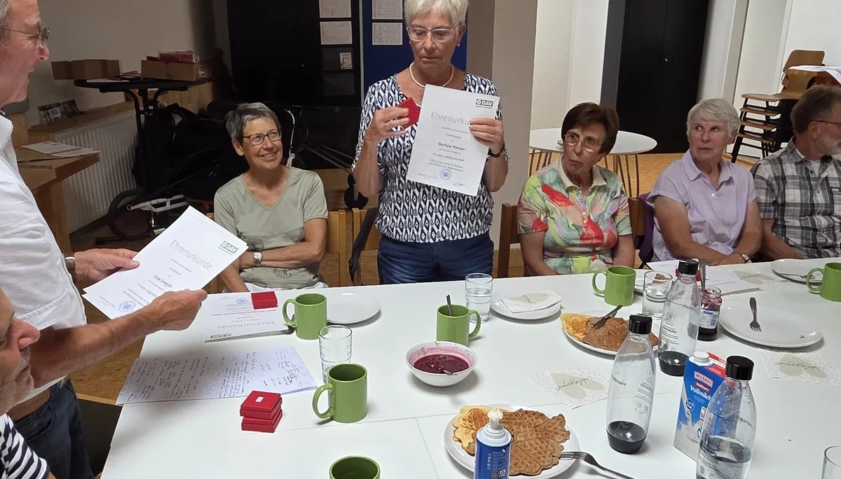 Eine Frau steht mit ihrer Ehrenurkunde und einer roten Schachtel in der Hand, während andere Mitglieder an einem großen Tisch sitzen und zuschauen. | © DAV Dortmund