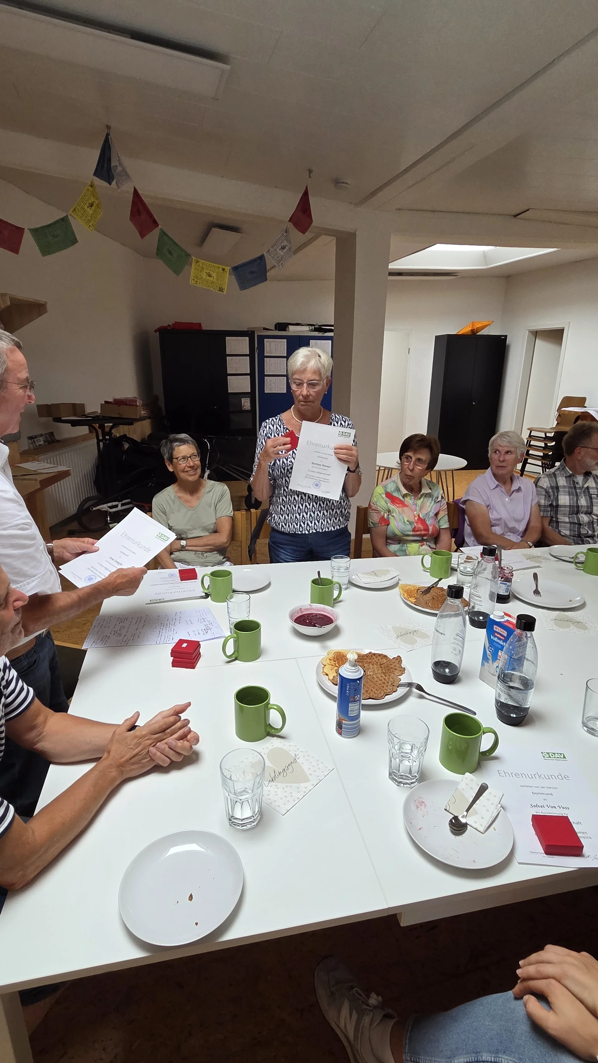 Eine Frau steht mit ihrer Ehrenurkunde und einer roten Schachtel in der Hand, während andere Mitglieder an einem großen Tisch sitzen und zuschauen. | © DAV Dortmund