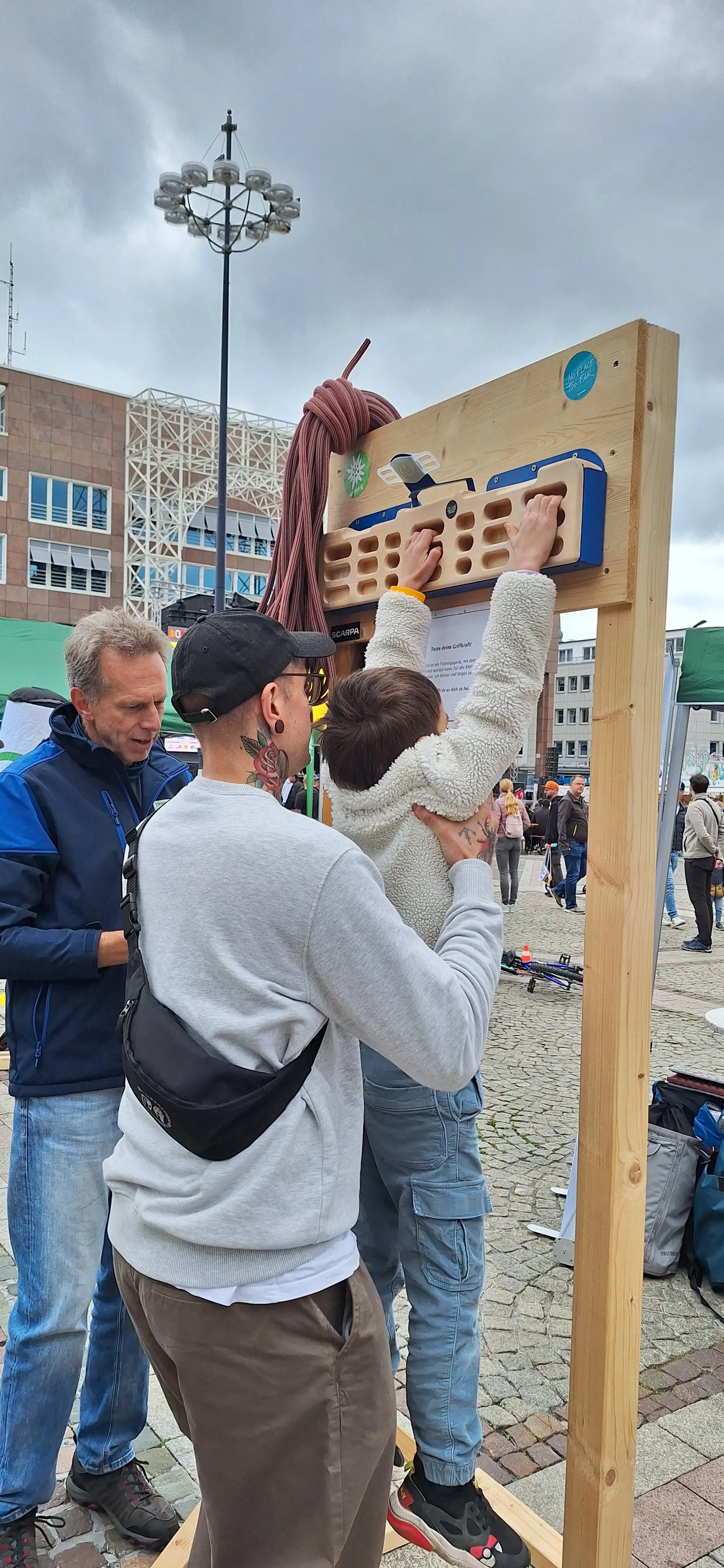 Ein Kind wird beim Hangeln an einem mobilen Trainingsboard auf einem Stadtplatz von zwei Erwachsenen unterstützt. | © DAV Dortmund