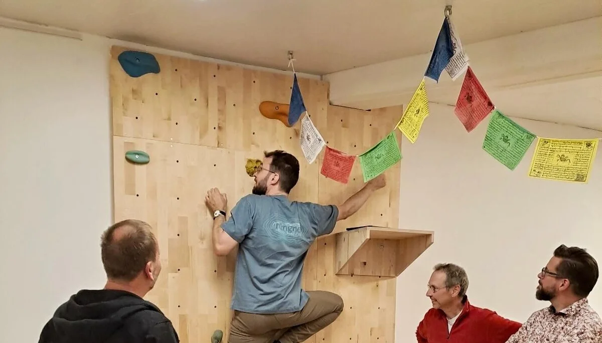 Eine Person klettert an einer Holzwand mit Griffen, umgeben von anderen Personen, die zuschauen. | © DAV Dortmund