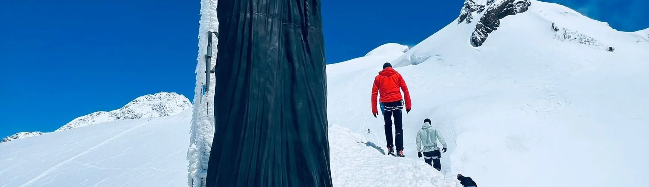 Eine dunkle Statue auf einem verschneiten Gipfel, dahinter Bergsteiger*innen bei sonnigem Wetter. | © DAV Dortmund