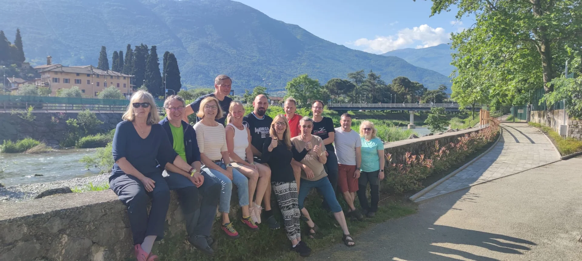 Gruppe von DAV-Mitgliedern sitzt lachend auf einer Mauer an einem Fluss mit Bergen im Hintergrund. | © DAV Dortmund