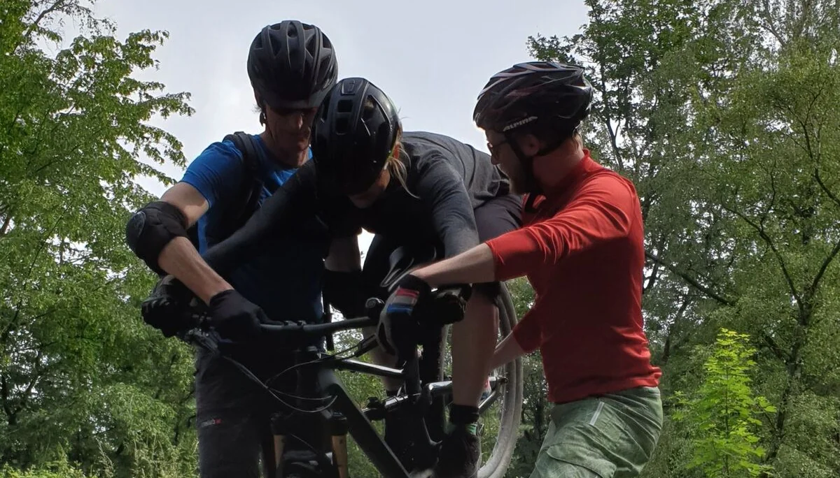 Drei Mountainbiker*innen stehen zusammen, einer auf dem Rad – technische Übungssituation. | © DAV Dortmund