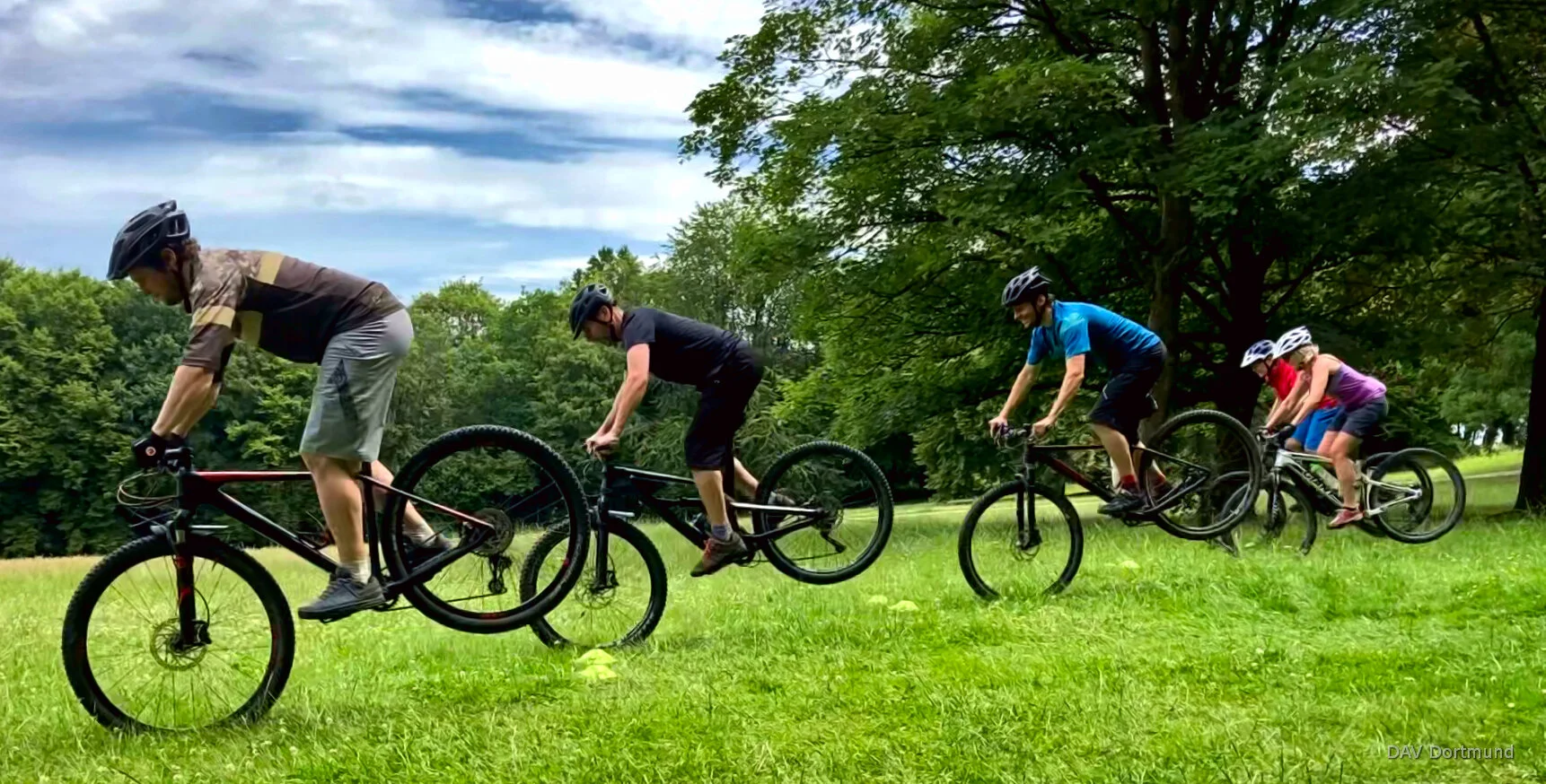 Vier Personen üben gleichzeitig eine Mountainbike-Technik auf einer Wiese mit Bäumen im Hintergrund. | © DAV Dortmund