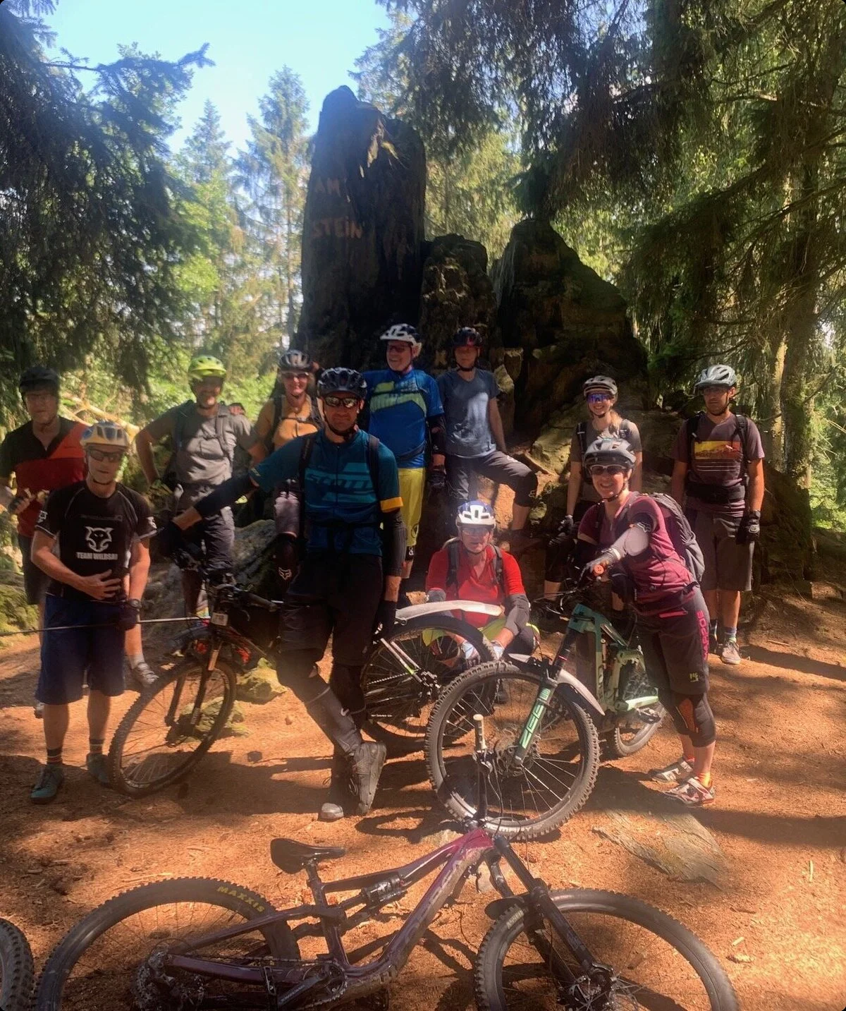 Mountainbike-Gruppe steht im Schatten großer Bäume bei einem Felsen im Wald. | © DAV Dortmund