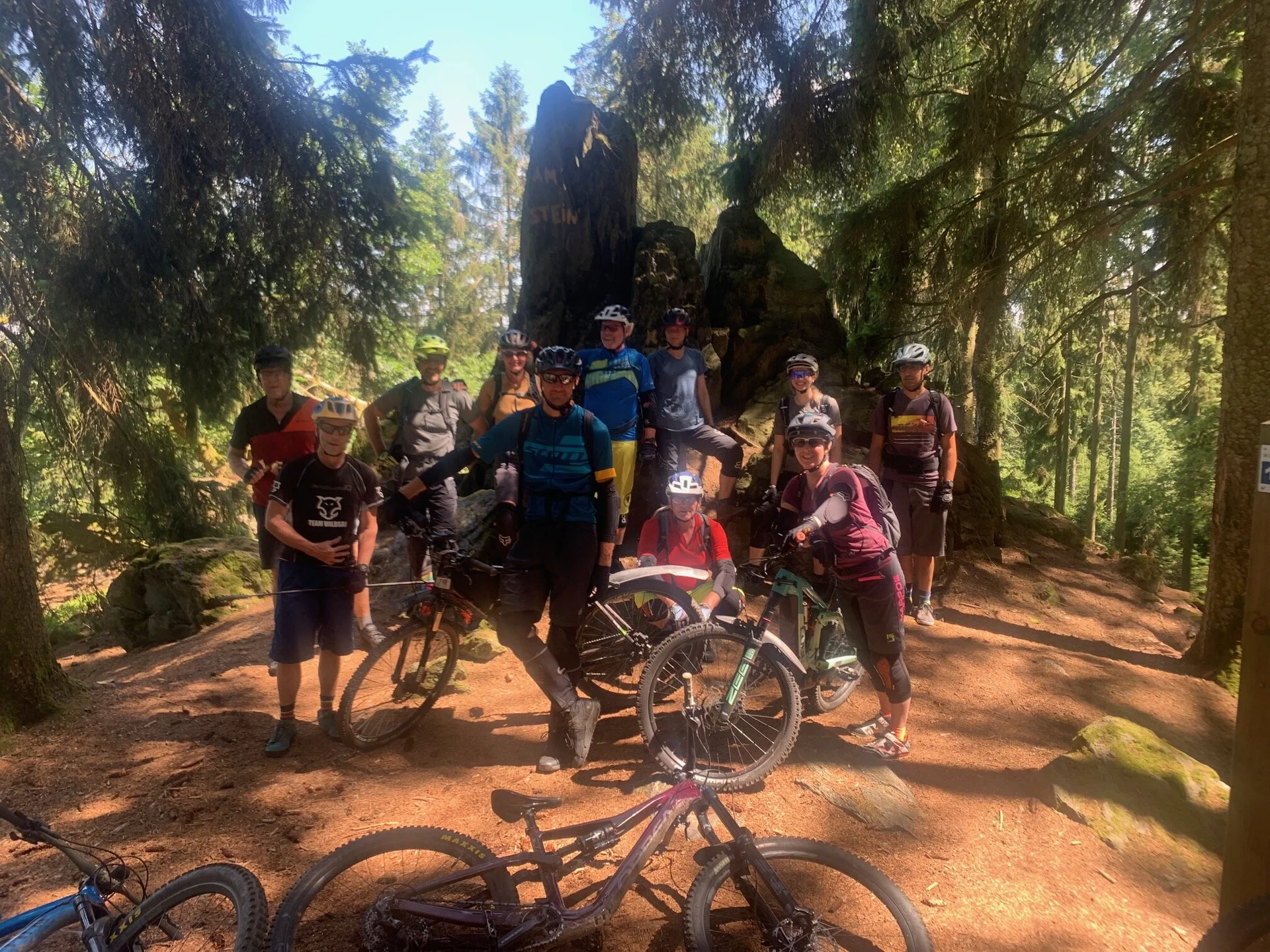 Mountainbike-Gruppe steht im Schatten großer Bäume bei einem Felsen im Wald. | © DAV Dortmund