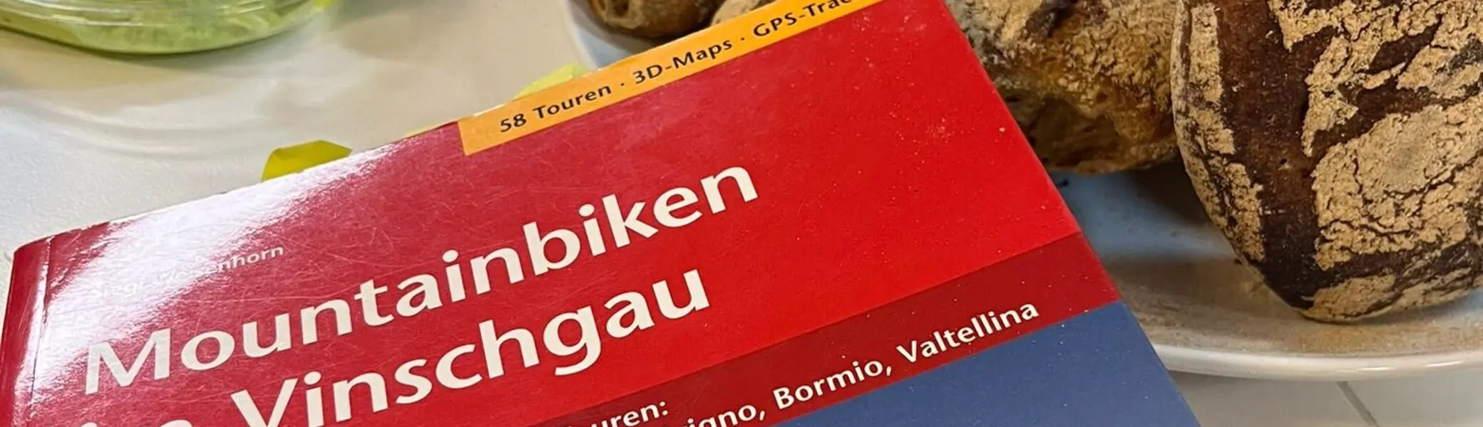 Nahaufnahme eines Reiseführers „Mountainbiken im Vinschgau“ auf einem Tisch mit Brot und Aufstrichen. | © dav mtb