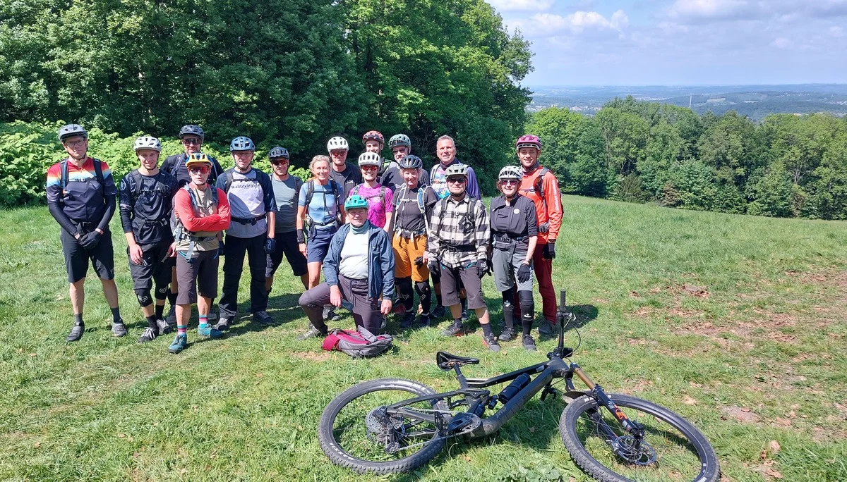Eine größere Gruppe von Mountainbiker*innen steht mit ihren Rädern auf einer Wiese vor Wald- und Talpanorama. | © DAV Dortmund
