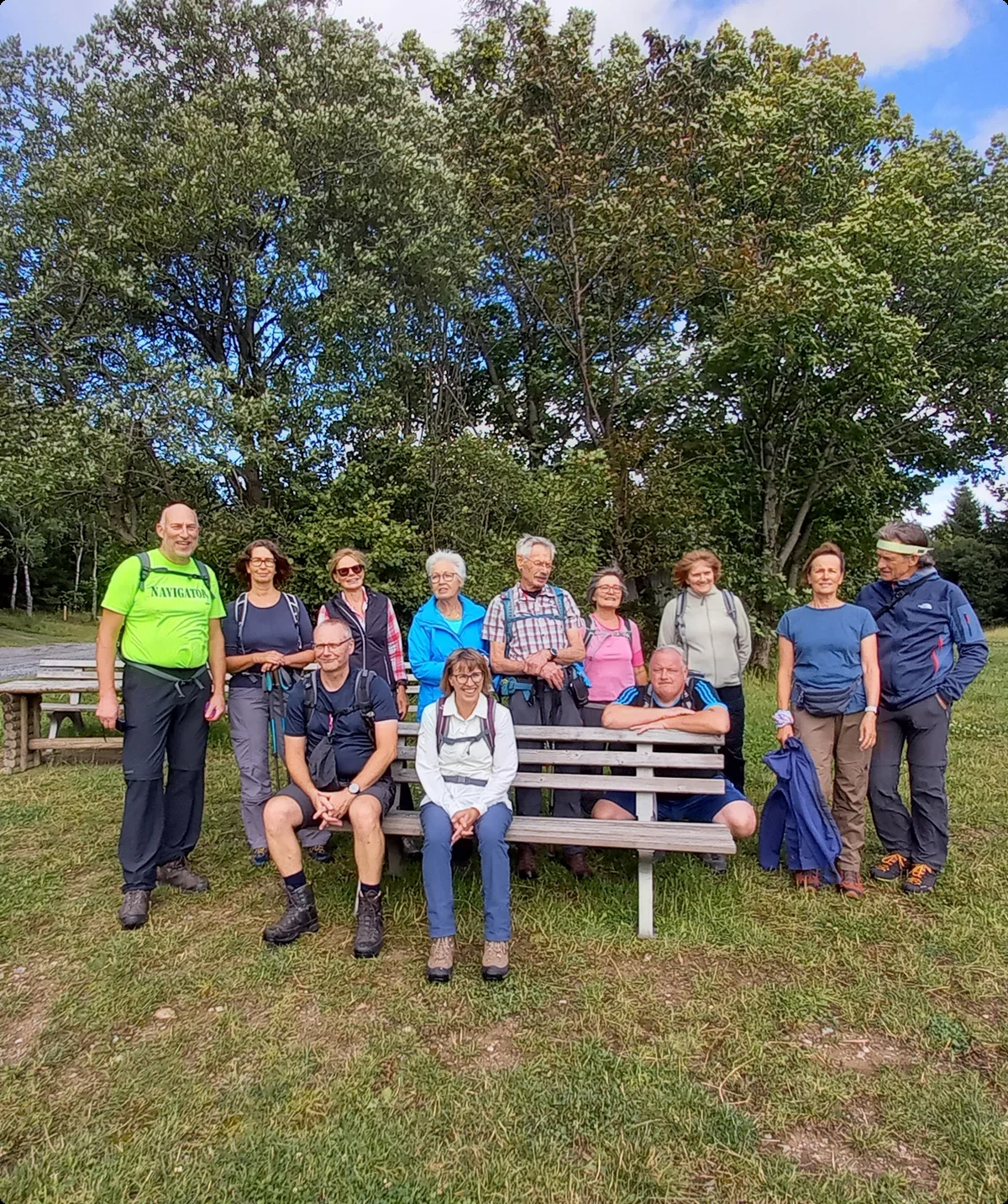 Eine Gruppe von Wandernden posiert auf einer Wiese neben Bänken unter Bäumen. | © DAV Dortmund