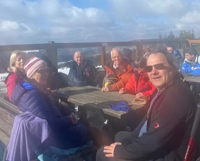 Wandergruppe sitzt bei sonnigem Wetter auf Holzbänken einer Terrasse im Schnee. | © DAV Dortmund