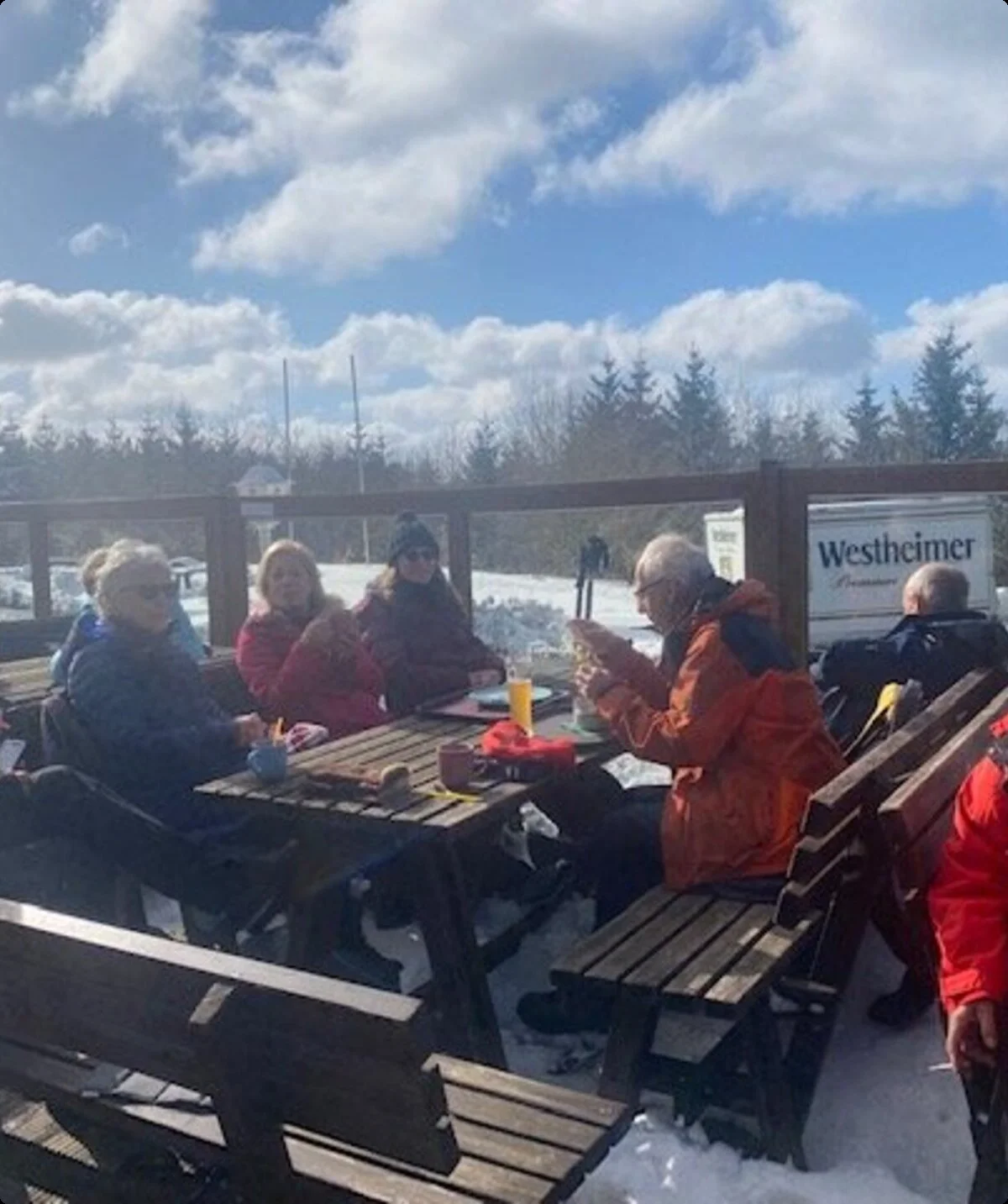 Wandergruppe sitzt bei sonnigem Wetter auf Holzbänken einer Terrasse im Schnee. | © DAV Dortmund