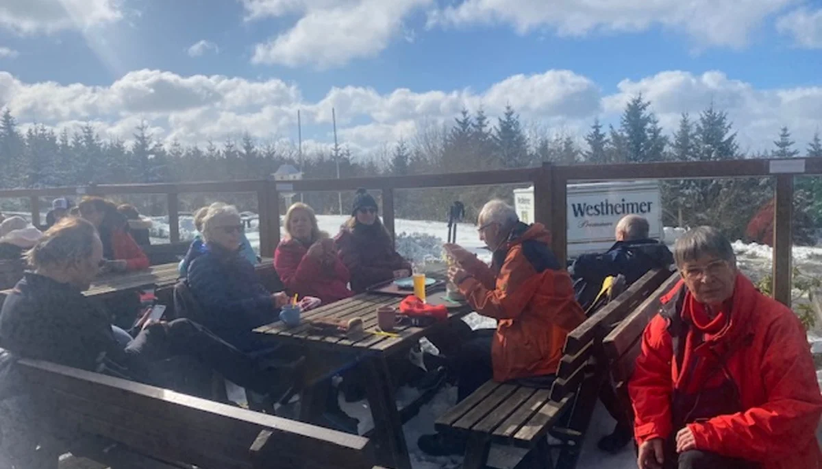 Wandergruppe sitzt bei sonnigem Wetter auf Holzbänken einer Terrasse im Schnee. | © DAV Dortmund