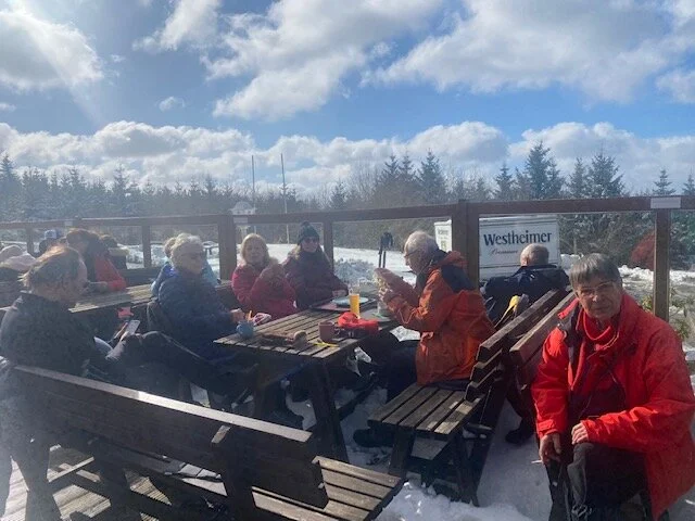 Wandergruppe sitzt bei sonnigem Wetter auf Holzbänken einer Terrasse im Schnee. | © DAV Dortmund