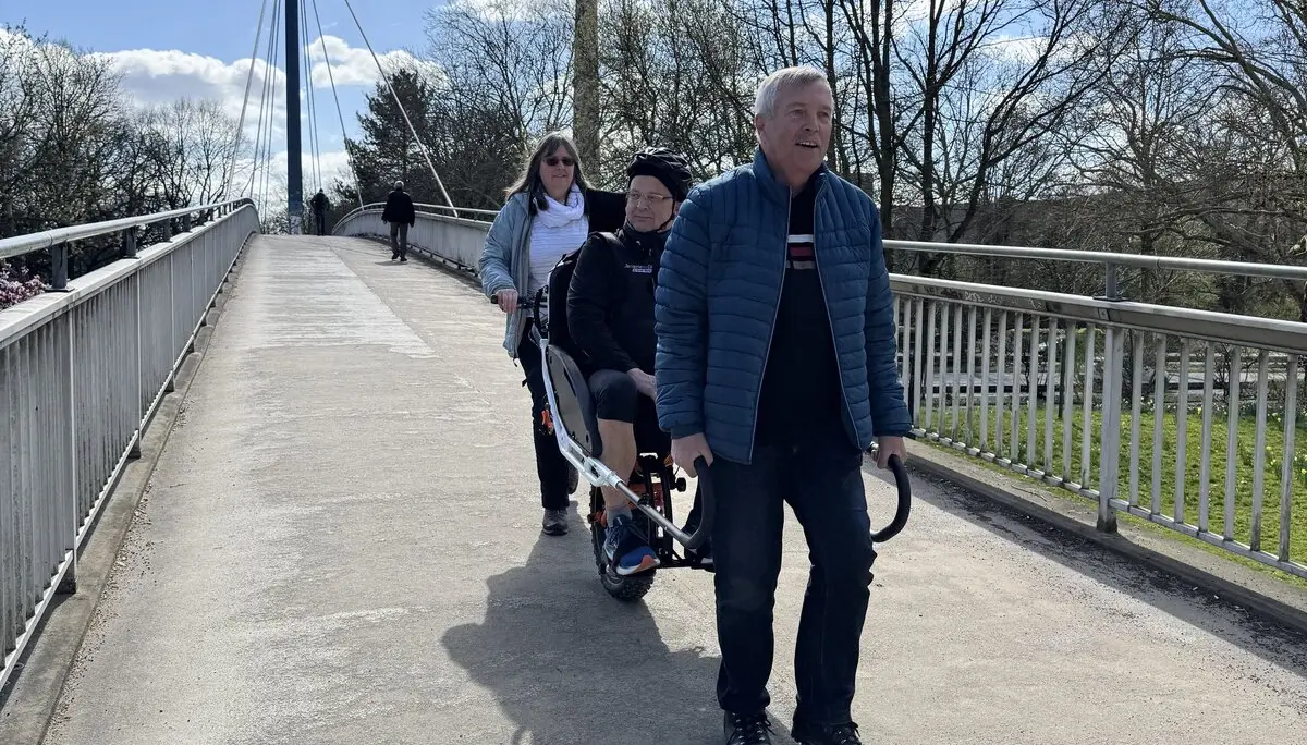 Drei Personen mit Spezialrollstuhl auf einer Brücke, Fernsehturm im Hintergrund. | © DAV Dortmund
