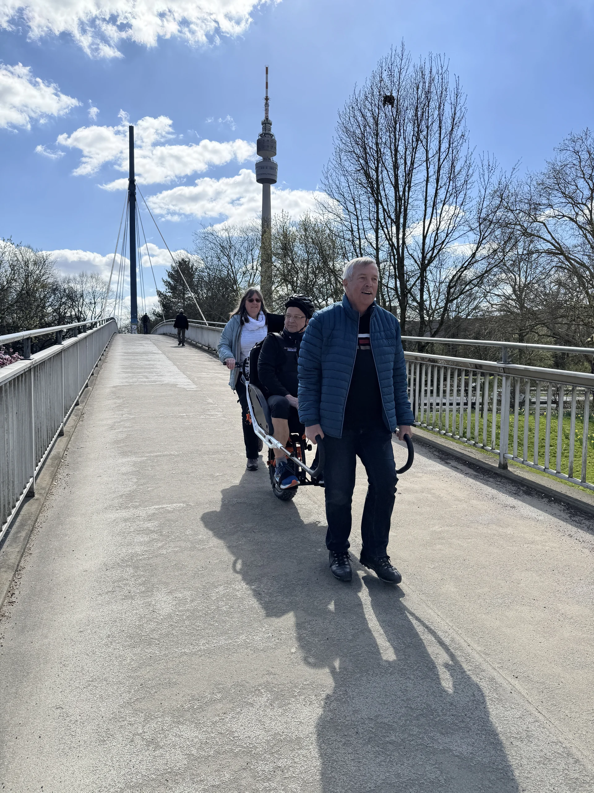 Drei Personen mit Spezialrollstuhl auf einer Brücke, Fernsehturm im Hintergrund. | © DAV Dortmund