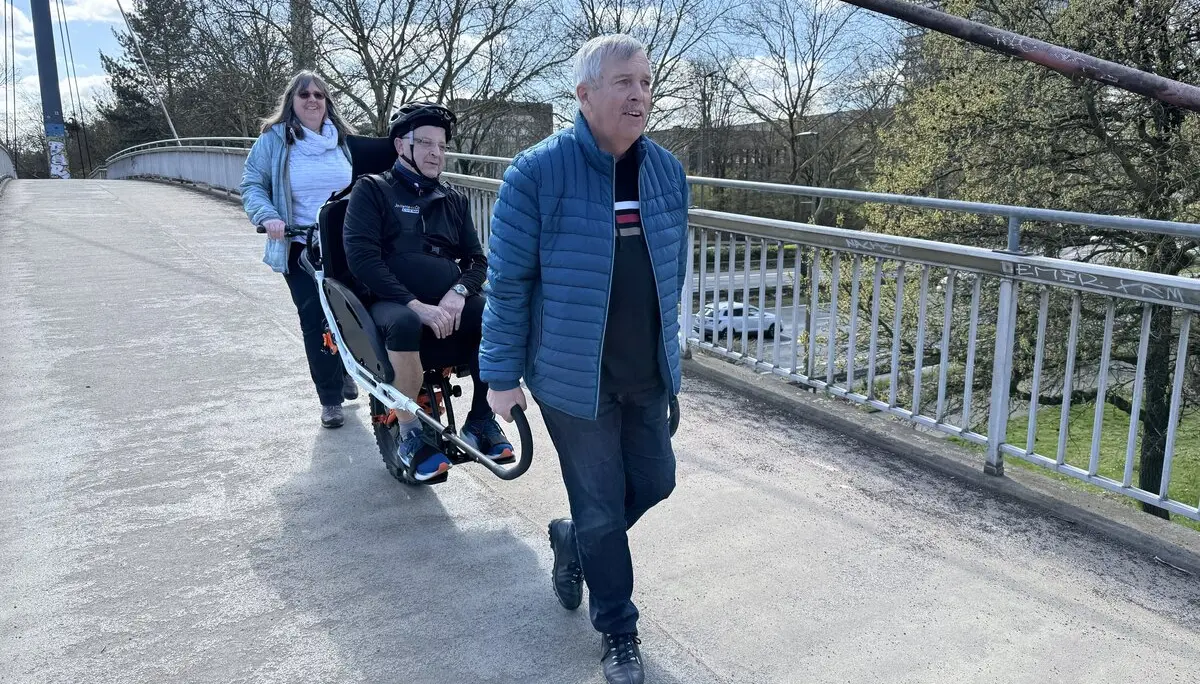 Drei Personen mit Spezialrollstuhl auf einer Brücke, Fernsehturm im Hintergrund. | © DAV Dortmund
