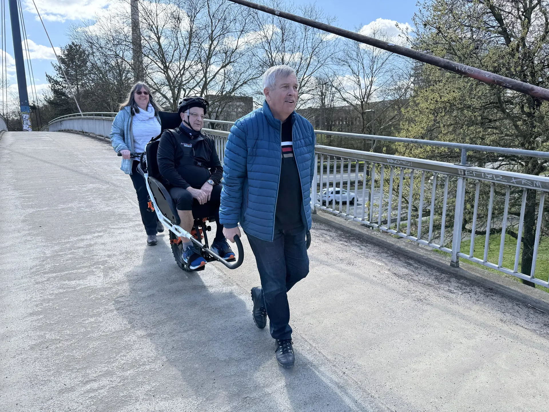Drei Personen mit Spezialrollstuhl auf einer Brücke, Fernsehturm im Hintergrund. | © DAV Dortmund