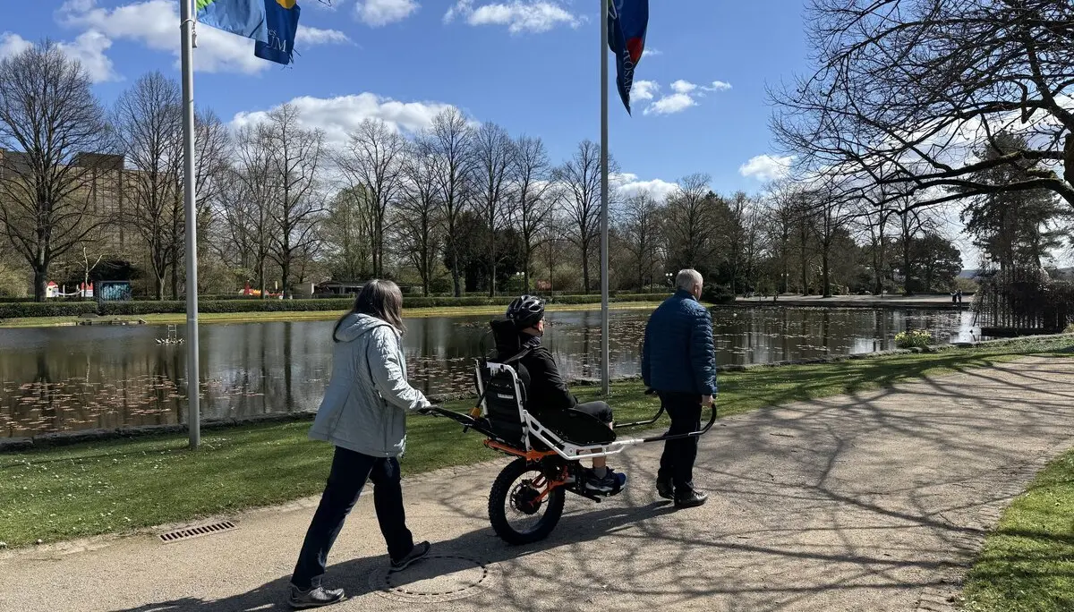 Person in einer Joëlette wird von zwei Begleitpersonen entlang eines Sees geschoben und gezogen. | © DAV Dortmund