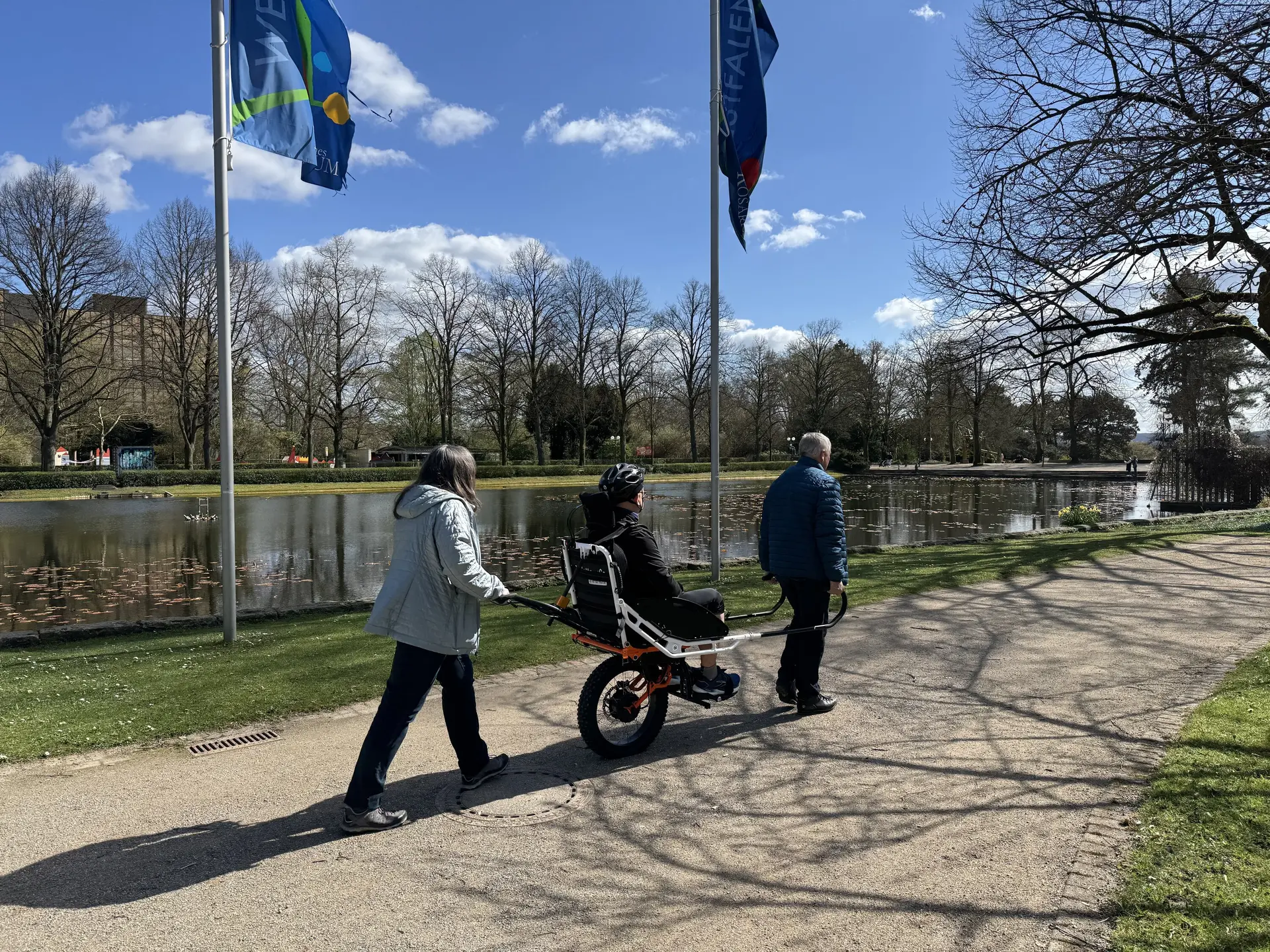 Person in einer Joëlette wird von zwei Begleitpersonen entlang eines Sees geschoben und gezogen. | © DAV Dortmund