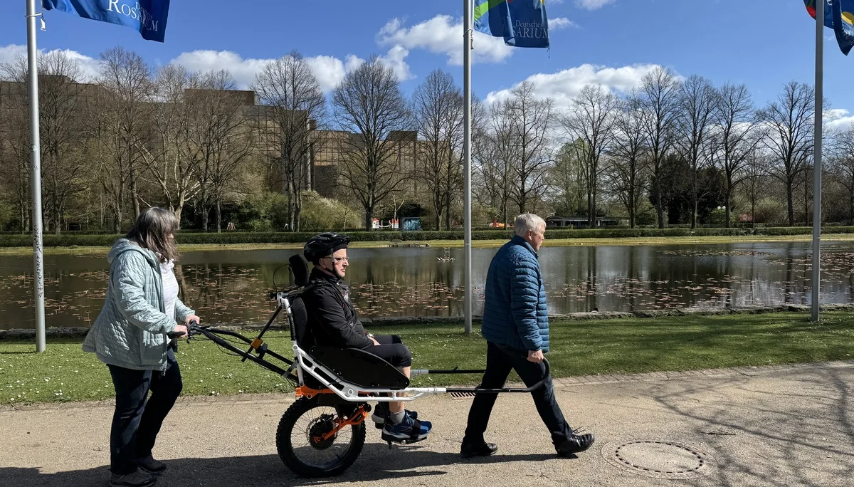 Person in einer Joëlette wird von zwei Begleitpersonen entlang eines Sees geschoben und gezogen. | © DAV Dortmund