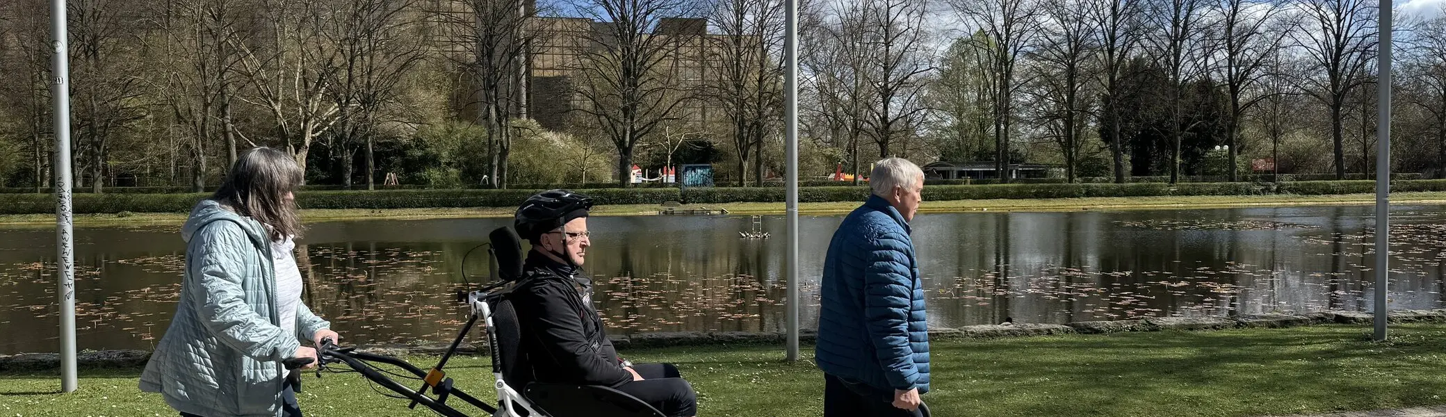 Person in einer Joëlette wird von zwei Begleitpersonen entlang eines Sees geschoben und gezogen. | © DAV Dortmund