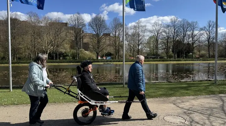 Person in einer Joëlette wird von zwei Begleitpersonen entlang eines Sees geschoben und gezogen. | © DAV Dortmund