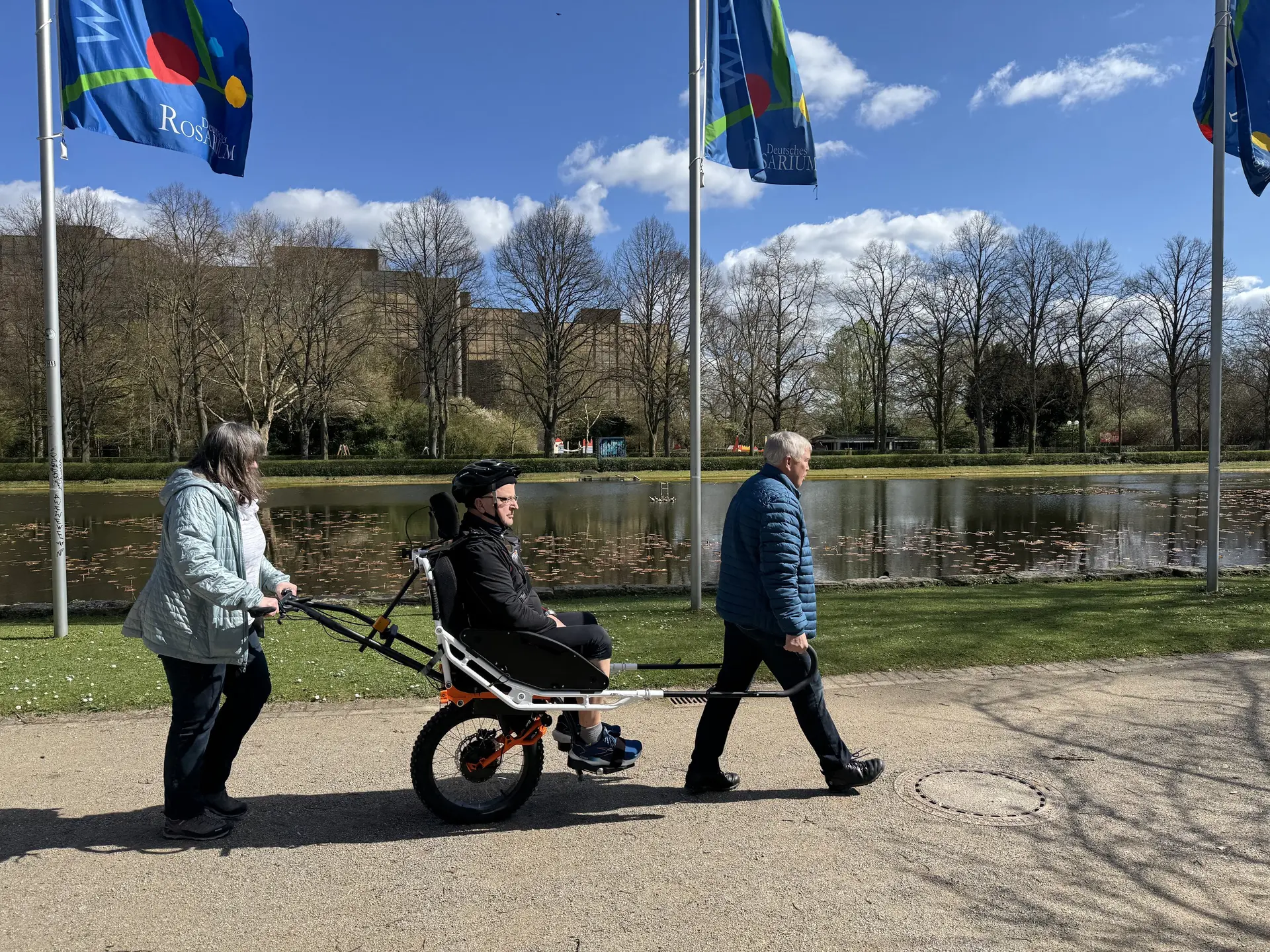 Person in einer Joëlette wird von zwei Begleitpersonen entlang eines Sees geschoben und gezogen. | © DAV Dortmund