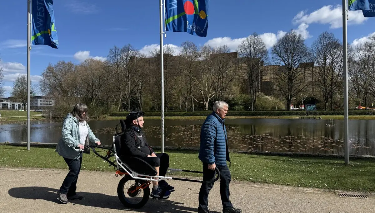 Person in einer Joëlette wird von zwei Begleitpersonen entlang eines Sees geschoben und gezogen. | © DAV Dortmund
