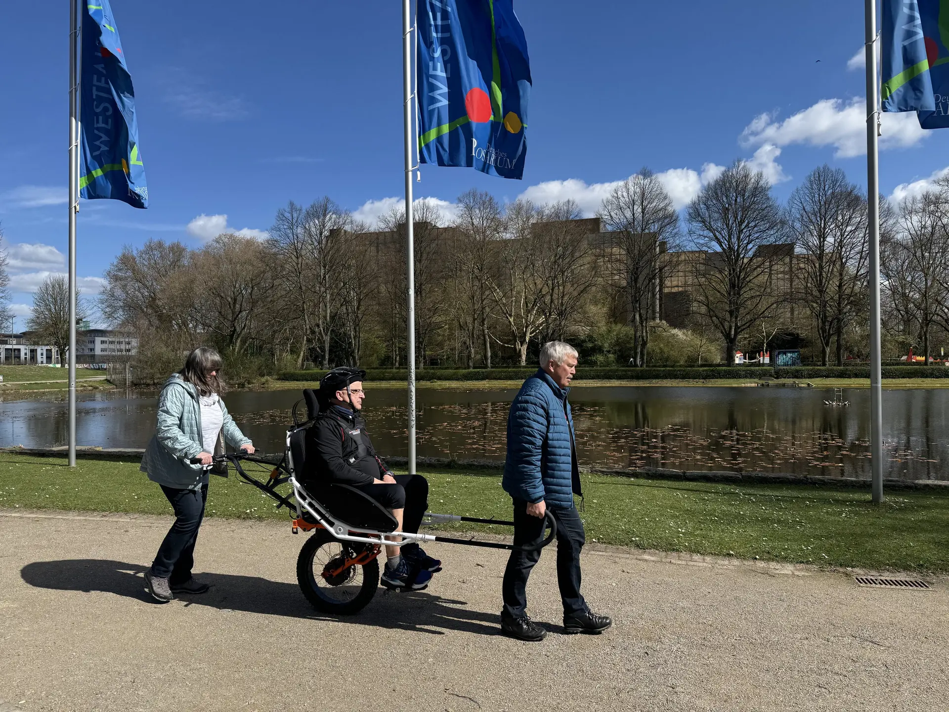 Person in einer Joëlette wird von zwei Begleitpersonen entlang eines Sees geschoben und gezogen. | © DAV Dortmund