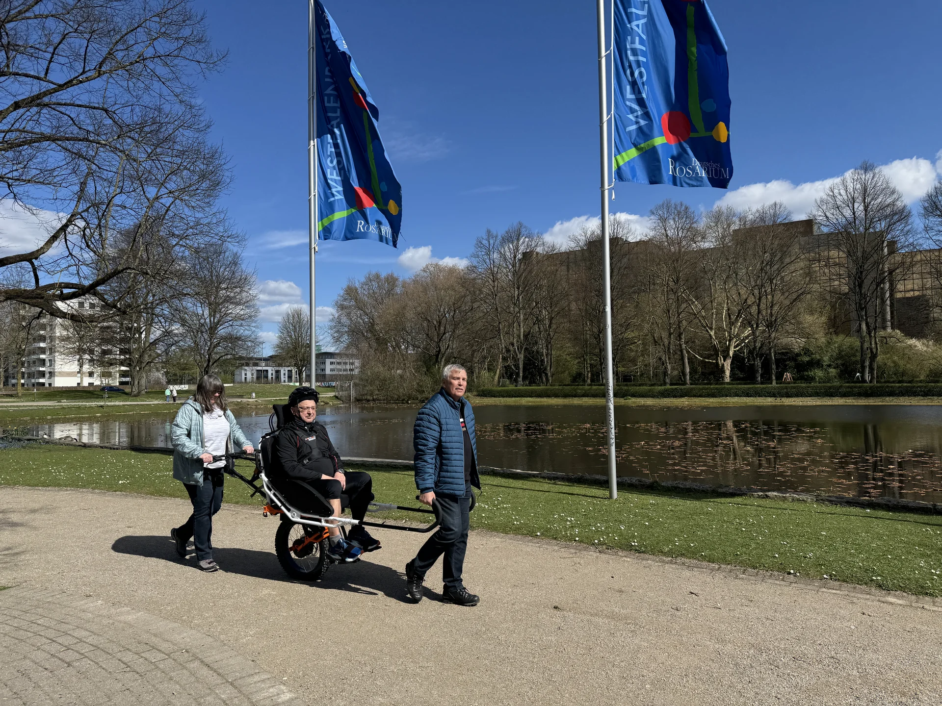 Drei Personen unterwegs mit einer Joëlette am Seeufer mit wehenden Fahnen. | © DAV Dortmund