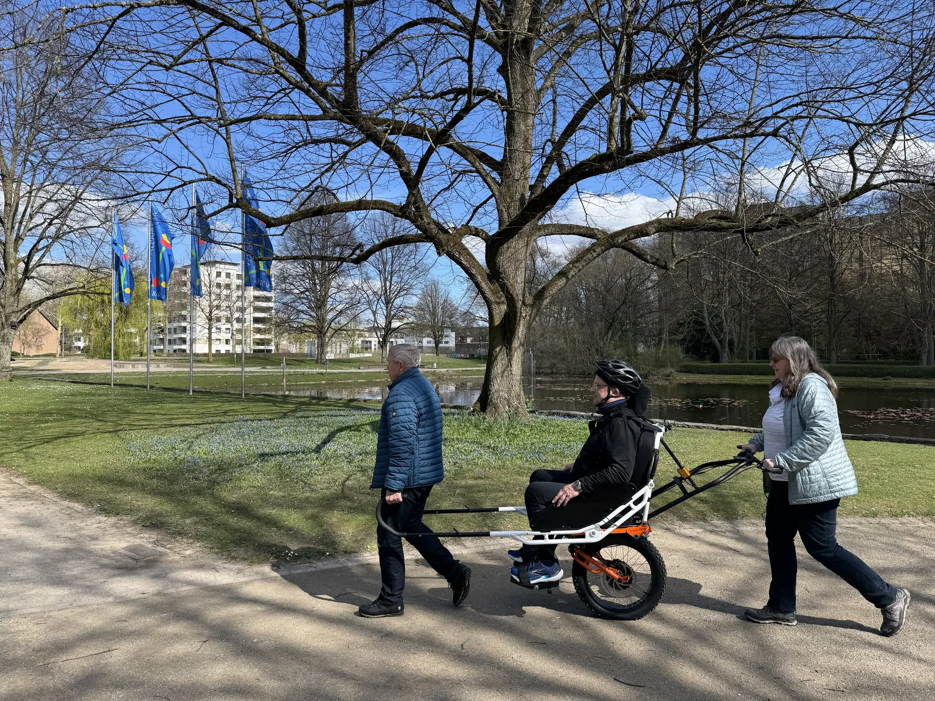 Person sitzt in einer geländegängigen Joëlette, zwei Begleitpersonen helfen beim Transport auf einem Parkweg. | © DAV Dortmund