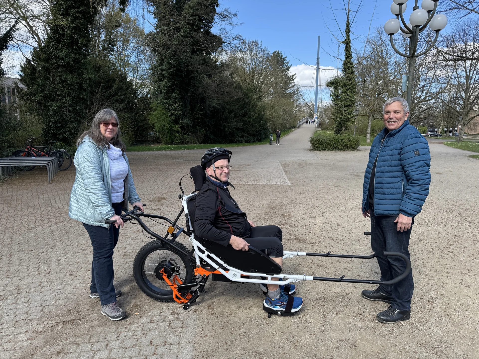 Person sitzt in einer geländegängigen Joëlette, zwei Begleitpersonen helfen beim Transport auf einem Parkweg. | © DAV Dortmund
