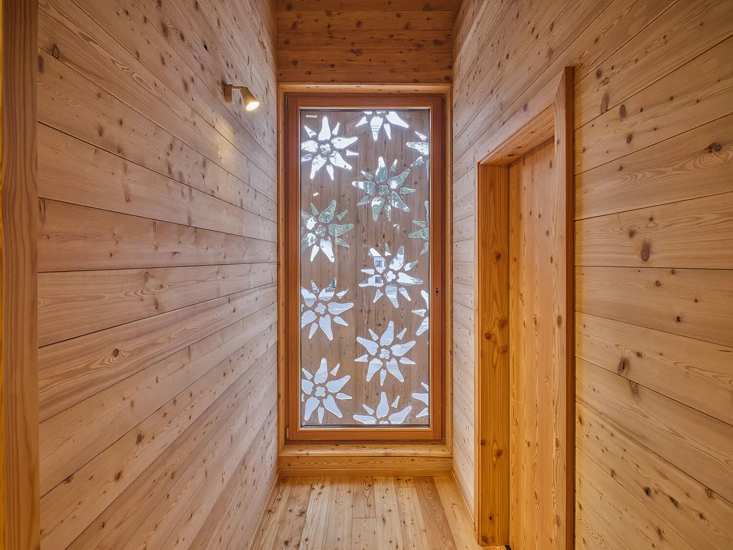 Großes Fenster mit Dekor aus Edelweißblüten am Ende eines Holzflurs in einer Berghütte. | © DAV Dortmund