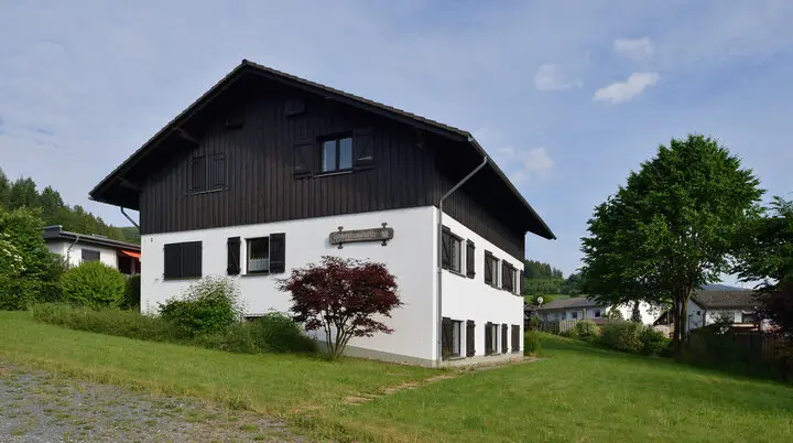 Eine zweistöckige Berghütte mit weißer Fassade und dunklem Holzaufsatz in grüner Umgebung. | © DAV Dortmund