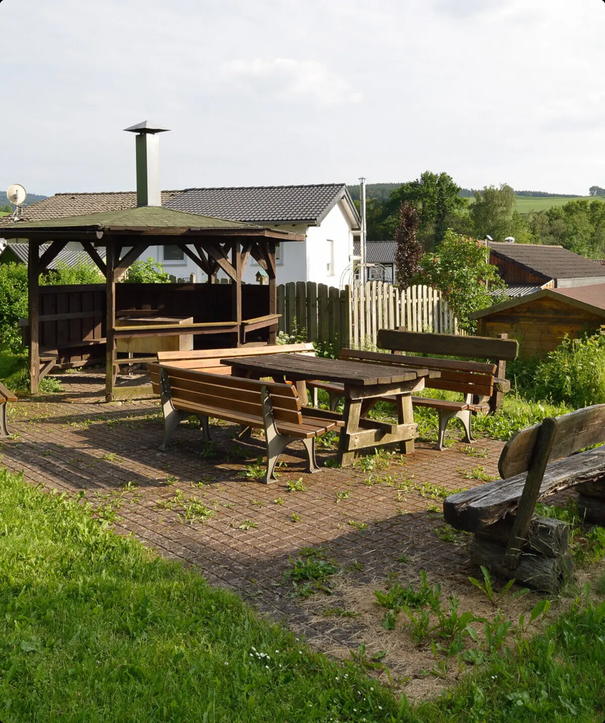 Ein gepflasterter Platz mit mehreren Holzbänken und einem überdachten Grillbereich neben einer Wiese. | © DAV Dortmund