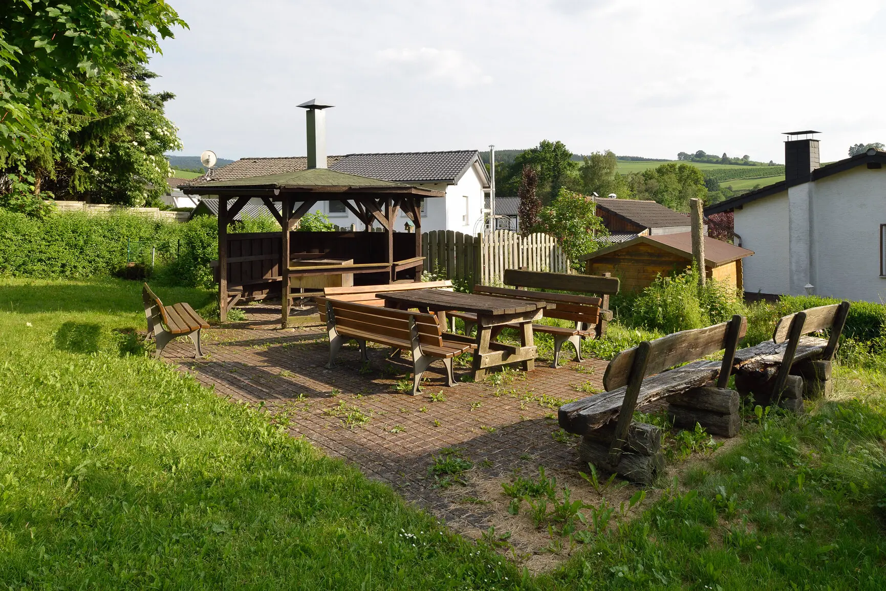 Ein gepflasterter Platz mit mehreren Holzbänken und einem überdachten Grillbereich neben einer Wiese. | © DAV Dortmund