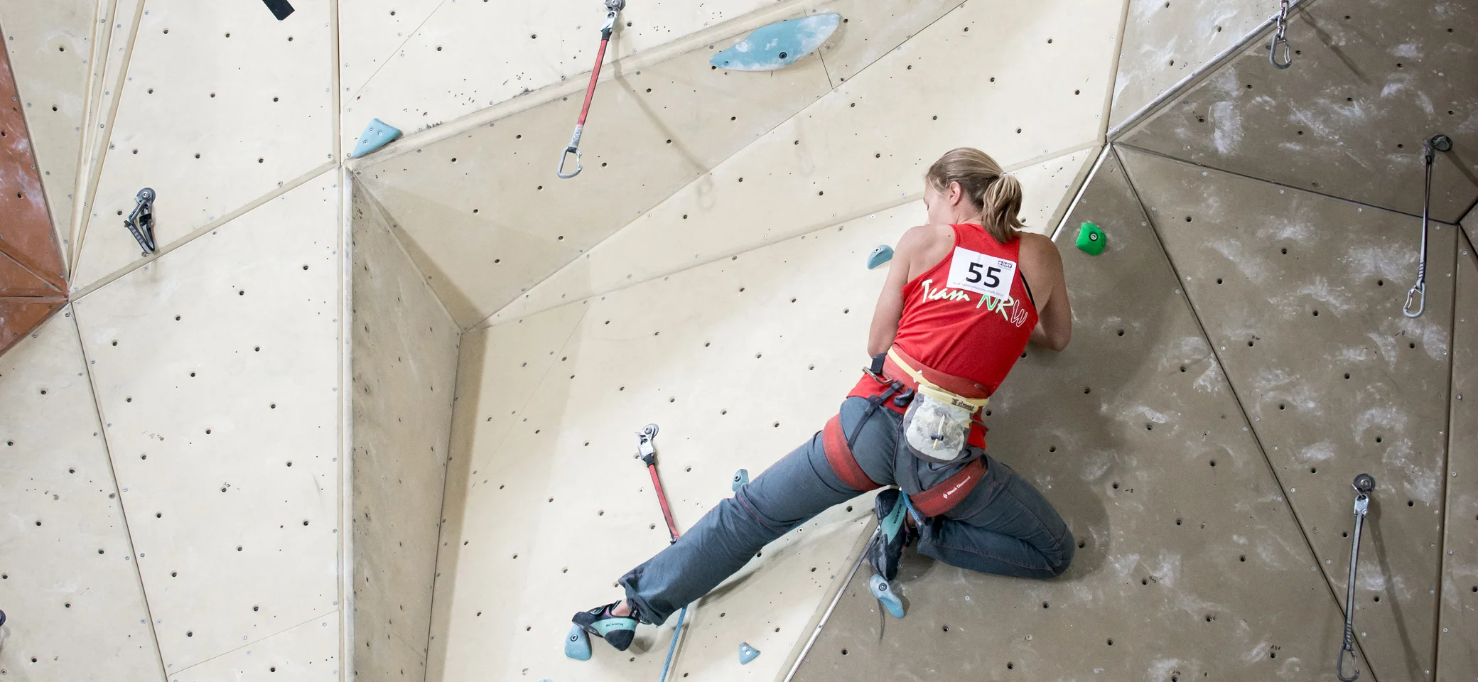 Person mit rotem Trikot klettert an einer strukturierten Indoor-Kletterwand mit Sicherungspunkten. | © DAV Dortmund