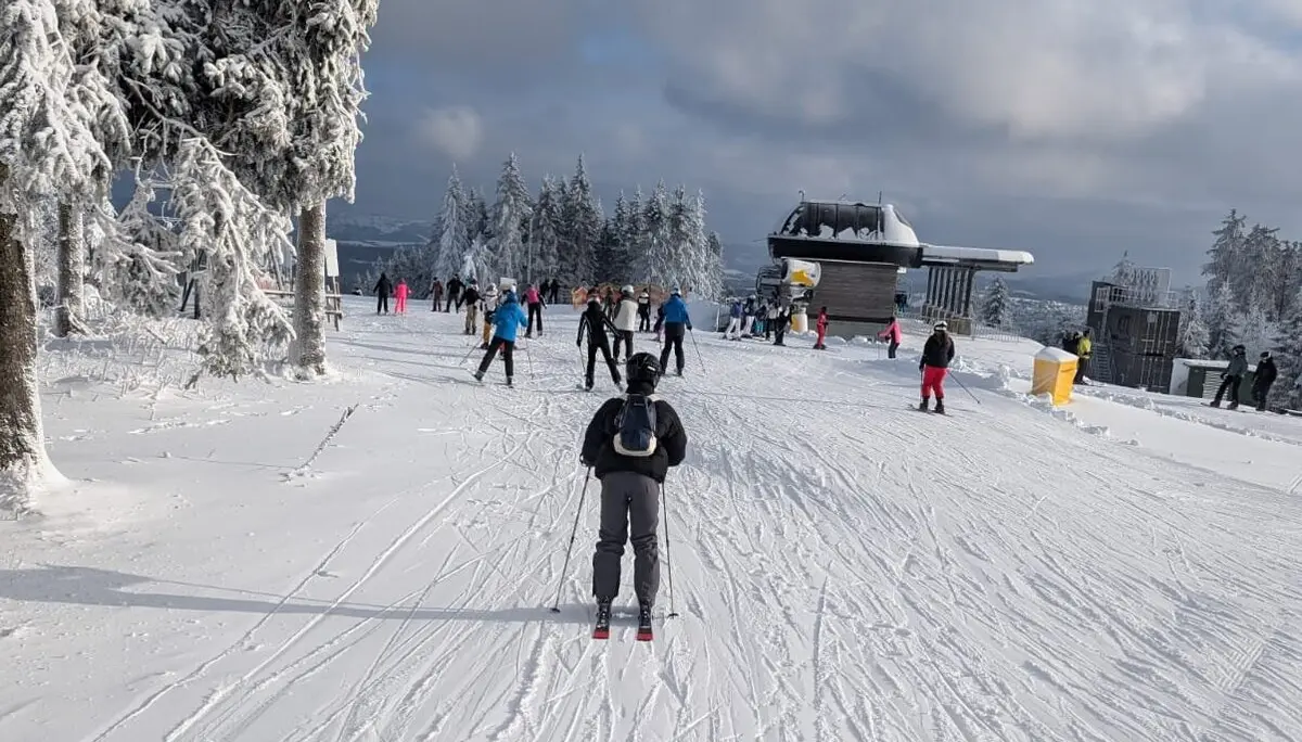 Einzelne und mehrere Skifahrer*innen auf breiter, sonniger Skipiste bei Bergstation. | © Dav Dortmund
